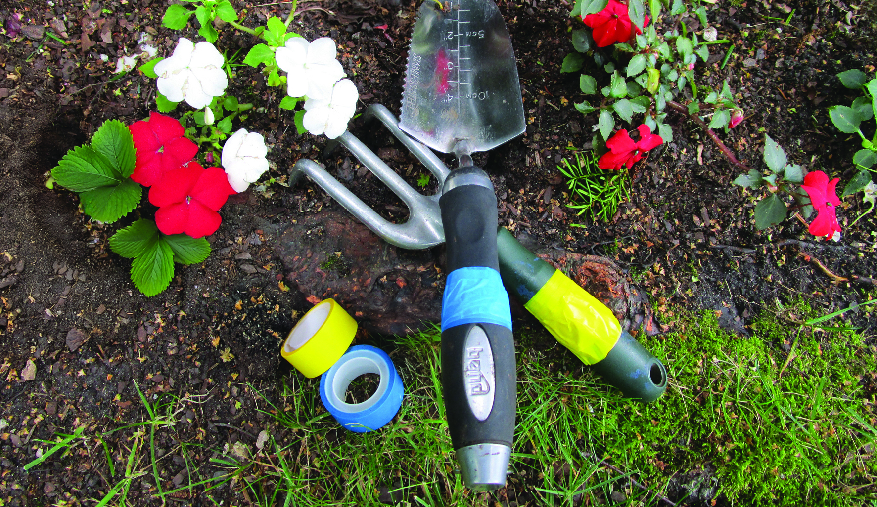 Two rolls of tape, yellow and blue, next to two garden hand tools with colored tape around their handles, on the dirt in a garden with some red and white impatiens.